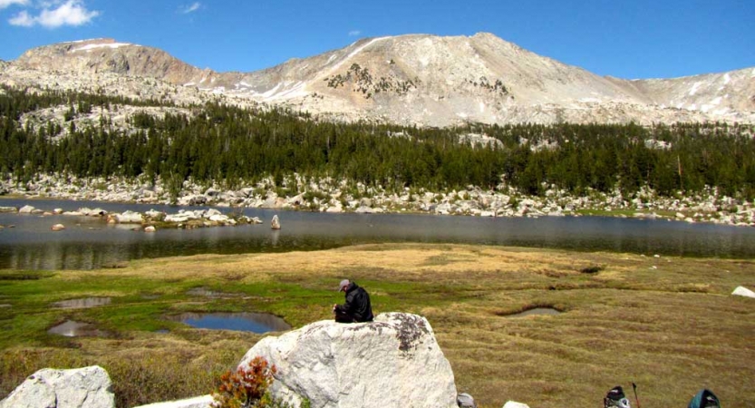 A person sits on a tall rock above a grassy landscape and appears to journal. There is a body of water, evergreen trees and mountains in the background.